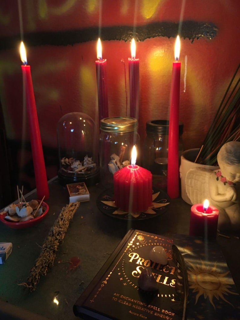 May include: A collection of red candles, including tall tapers and a thick pillar candle, arranged with a book titled Protection Spells, a glass jar, a small glass dome, a white angel figurine, and a bundle of dried herbs on a dark surface.