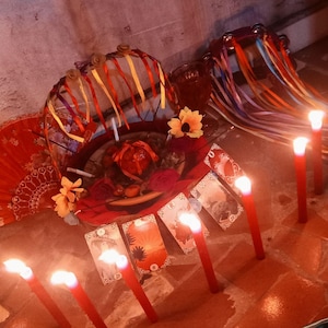 May include: A collection of items including a basket decorated with colorful ribbons and artificial flowers, a red fan, a tambourine with ribbons, and several tarot cards arranged in a row, all illuminated by multiple lit red candles.
