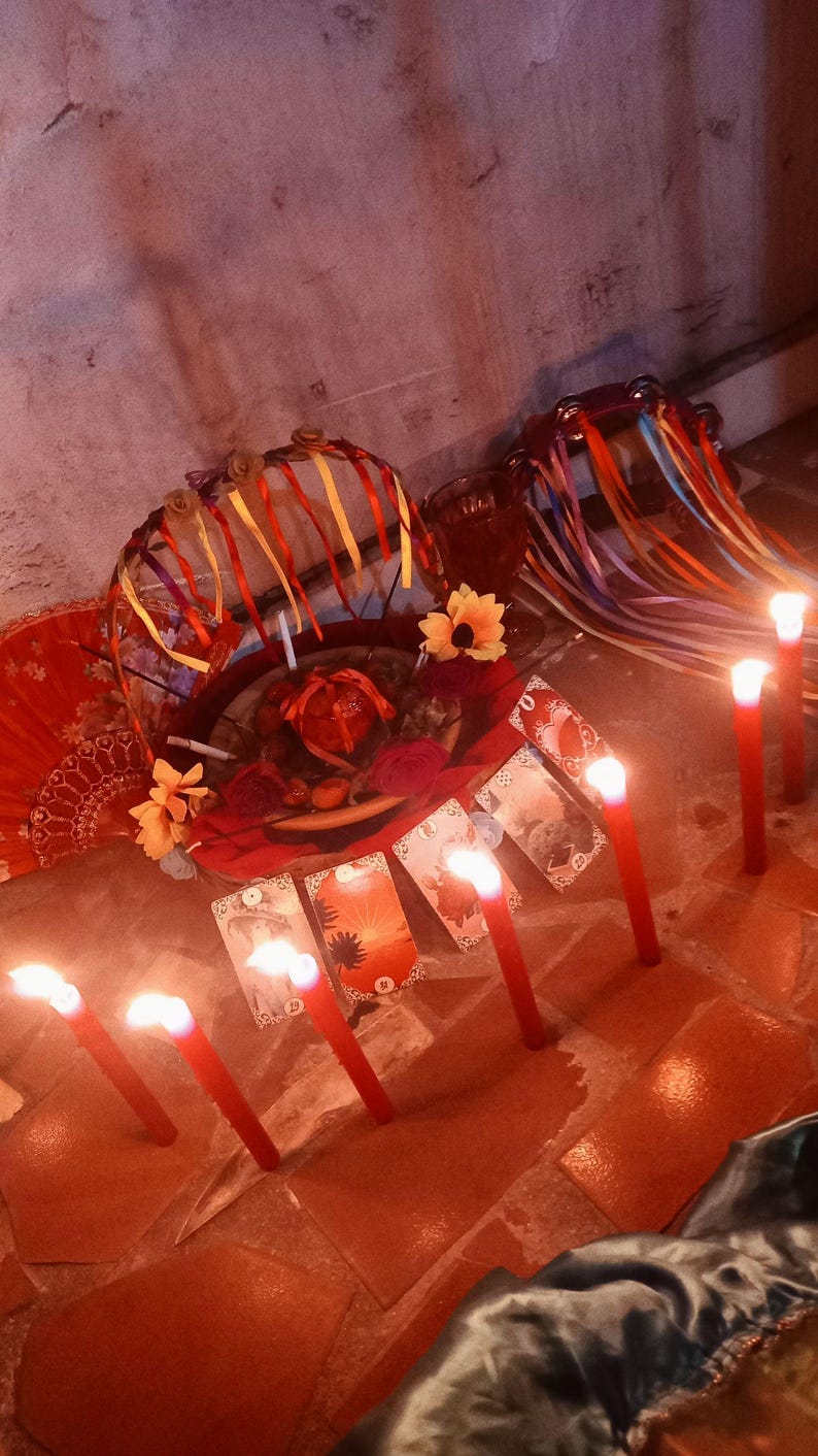 May include: A collection of items including a basket decorated with colorful ribbons and artificial flowers, a red fan, a tambourine with ribbons, and several tarot cards arranged in a row, all illuminated by multiple lit red candles.