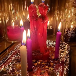 Puede incluir: Dos velas rojas con forma de figura est&aacute;n juntas, cubiertas de purpurina, rodeadas de velas c&oacute;nicas m&aacute;s peque&ntilde;as de color morado y blanco. Est&aacute;n sobre una base de papel de aluminio con p&eacute;talos secos y portavelas de cristal al fondo.