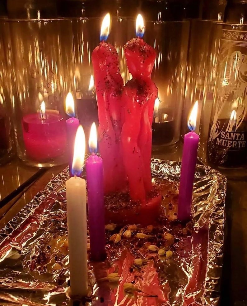 Puede incluir: Dos velas rojas con forma de figura est&aacute;n juntas, cubiertas de purpurina, rodeadas de velas c&oacute;nicas m&aacute;s peque&ntilde;as de color morado y blanco. Est&aacute;n sobre una base de papel de aluminio con p&eacute;talos secos y portavelas de cristal al fondo.