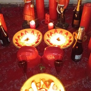 May include: A red lace tablecloth holds two ceramic bowls filled with sliced strawberries and cream, a third bowl with fruit, two tall red candles, two smaller red candles, two figurines, two wine glasses, and two bottles of champagne.