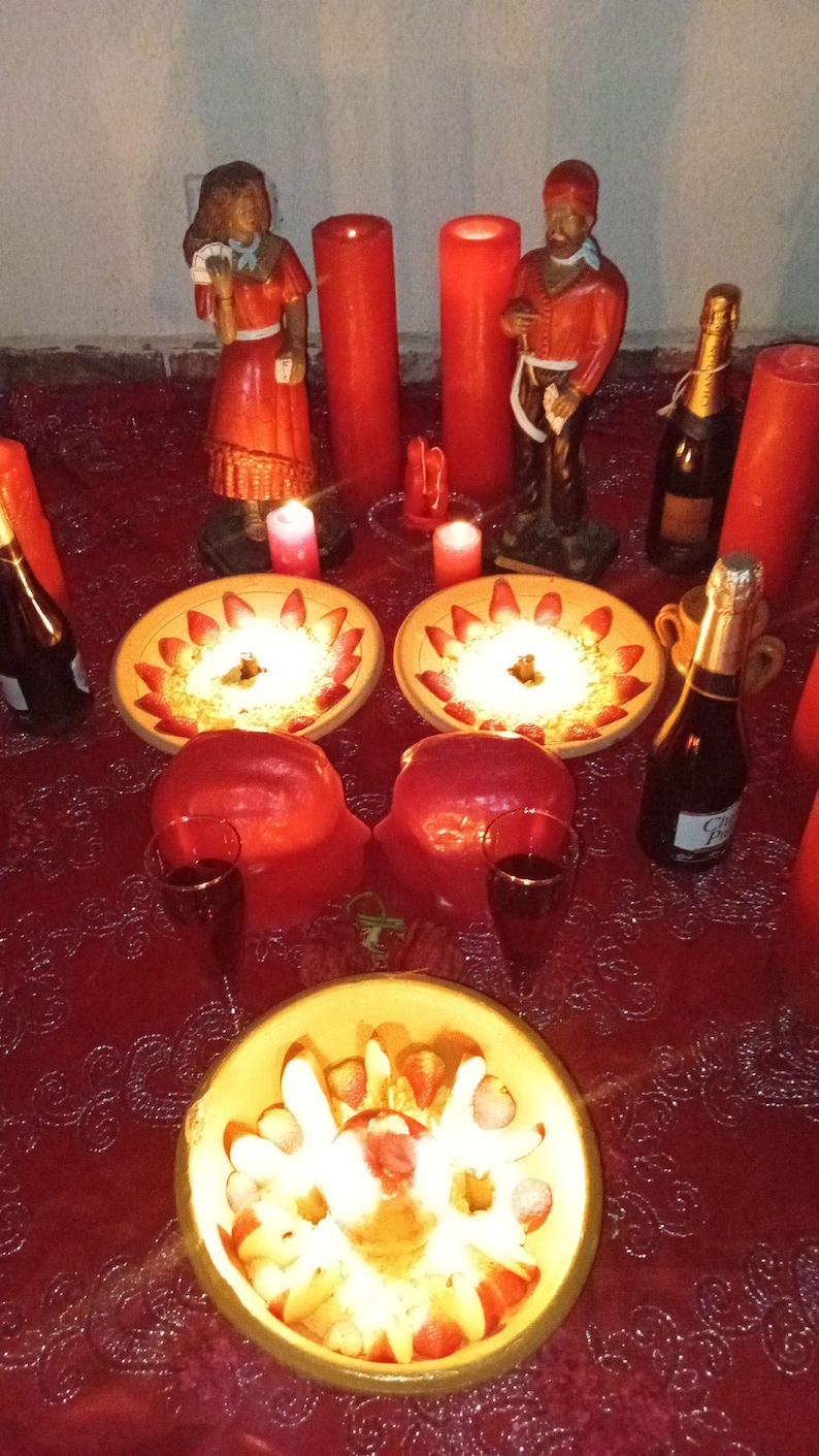 May include: A red lace tablecloth holds two ceramic bowls filled with sliced strawberries and cream, a third bowl with fruit, two tall red candles, two smaller red candles, two figurines, two wine glasses, and two bottles of champagne.