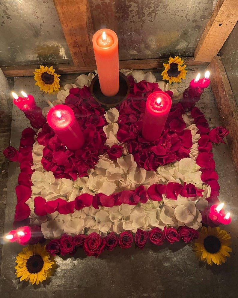 May include: A square arrangement of white and deep red rose petals, bordered by red roses and four yellow sunflowers at the corners. Five lit pillar candles in shades of orange and red are placed within the floral display, casting a warm glow.