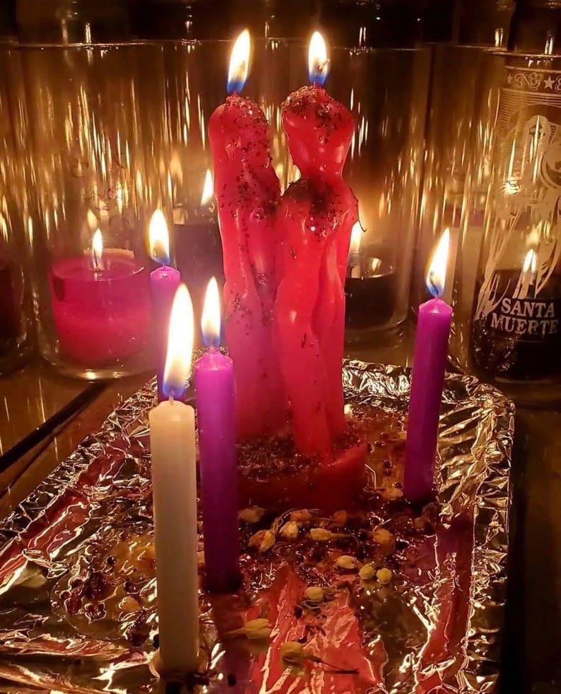 May include: Two red figure candles stand together, topped with glitter, surrounded by smaller purple and white taper candles. The candles are arranged on a foil-covered base with scattered dried flower petals, with glass candle holders in the background.
