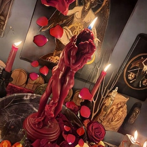 May include: A deep red wax candle sculpted into the shape of two figures embracing, standing on a circular base. The candle is lit, surrounded by scattered red rose petals, additional lit taper candles, and various decorative objects.