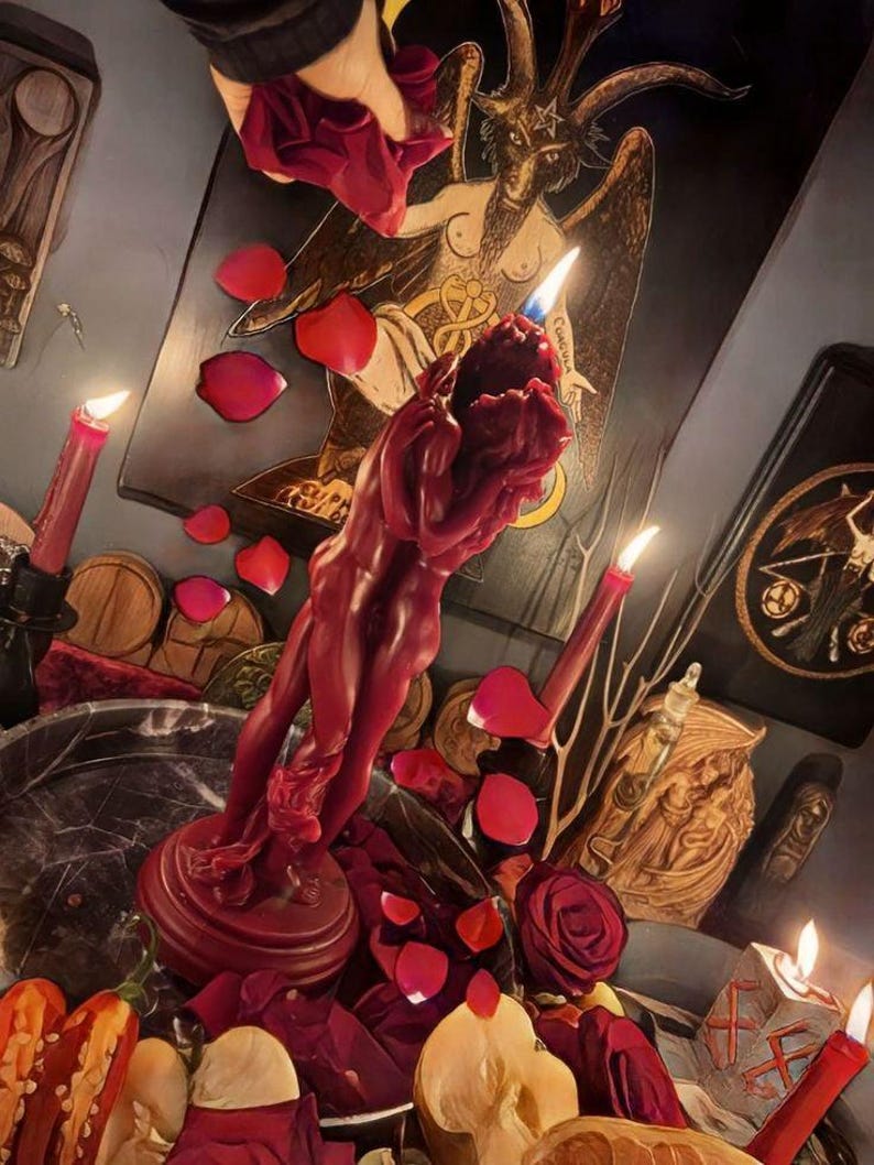 May include: A deep red wax candle sculpted into the shape of two figures embracing, standing on a circular base. The candle is lit, surrounded by scattered red rose petals, additional lit taper candles, and various decorative objects.
