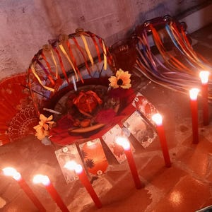 Puede incluir: Una colecci&oacute;n de objetos que incluye una cesta decorada con cintas de colores y flores artificiales, un abanico rojo, una pandereta con cintas y varias cartas del tarot dispuestas en fila, todo iluminado por varias velas rojas encendidas.