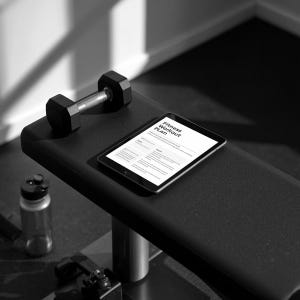 Può includere: Un tablet nero che mostra un documento Fitness Workout Plan, appoggiato su una panca da allenamento nera accanto a un manubrio nero. Lo schermo mostra testo tra cui Fitness Workout Plan, Goals e Results, con un layout chiaro per il monitoraggio.