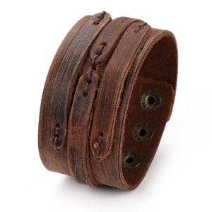 Puede incluir: Una pulsera de cuero marrón ancha con textura desgastada y diseño en capas. La pulsera está construida con múltiples tiras de cuero unidas por costuras de color marrón oscuro e incluye tres botones de presión metálicos para el cierre.