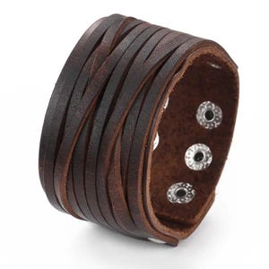 Op de afbeelding: Een brede donkerbruine leren manchetarmband bestaande uit meerdere dunne, overlappende leren stroken. De armband is voorzien van een reeks zilverkleurige metalen drukknopen aan de binnenzijde om de band rond de pols te bevestigen.