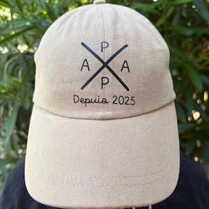 Peut inclure: Une casquette de baseball beige avec un motif graphique noir composé de deux lignes croisées et des lettres P, A, P et A placées dans les quatre quadrants. Sous les lignes croisées, le texte "Depuis 2025" est imprimé en police cursive noire.