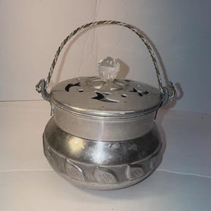 May include: A silver-toned metal pot with a rounded body featuring a band of embossed leaf patterns. It includes a removable lid with cut-out shapes and a clear crystal-like knob, plus a twisted metal wire handle attached to the sides.