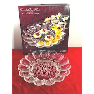 May include: A clear glass deviled egg plate with a textured, scalloped edge and a central circular pattern. The plate is displayed in front of its original dark red box, which features the text Deviled Egg Plate and Plateau a Hors d'oeuvre Pour Oeufs.