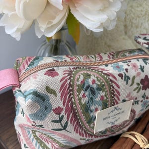 Puede incluir: Un estuche rectangular de tela con fondo crema y un estampado floral y de cachemira multicolor en tonos rosa, verde y azul. Tiene una cremallera de color cobre, una lengüeta lateral rosa y una etiqueta blanca con el texto ROSE-CANNELLE.
