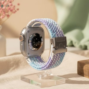 Może przedstawiać: Srebrny smartwatch z okrągłym czujnikiem z tyłu, przymocowany do plecionego elastycznego paska do zegarka z gradientowym wzorem w kolorze jasnoniebieskim, lawendowym i białym. Pasek posiada srebrną metalową klamrę.
