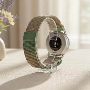 May include: A smartwatch with a circular silver casing and a two-tone woven mesh band in beige and sage green. The watch is displayed on a clear acrylic stand, showing the back of the device with its integrated circular sensor array and metallic finish.