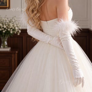 May include: A strapless white gown featuring a full tulle skirt and long satin gloves. The gloves are adorned with white feather trim at the upper arms. The text Dainty Details is visible in the upper left and lower right corners of the image.
