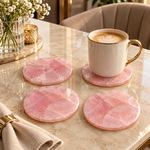 May include: A set of four circular rose quartz coasters with a polished finish, showcasing natural pink crystalline patterns. One coaster supports a white ceramic mug filled with coffee, while the other three are arranged on a flat surface.