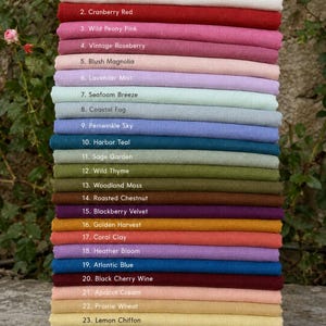 Op de afbeelding: Een stapel van vijfentwintig gevouwen stoffen stalen in verschillende kleuren, elk gelabeld met een nummer en naam, waaronder Alpine Snow, Cranberry Red, Wild Peony Pink, Vintage Roseberry, Blush Magnolia, Lavender Mist, Seafoam Breeze, Coastal Fog, Periwinkle Sky, Harbor Teal, Sage Garden, Wild Thyme, Woodland Moss, Roasted Chestnut, Blackberry Velvet, Golden Harvest, Coral Clay, Heather Bloom, Atlantic Blue, Black Cherry Wine, Apricot Cream, Prairie Wheat, Lemon Chiffon, Pine Grove en Canyon Oak.