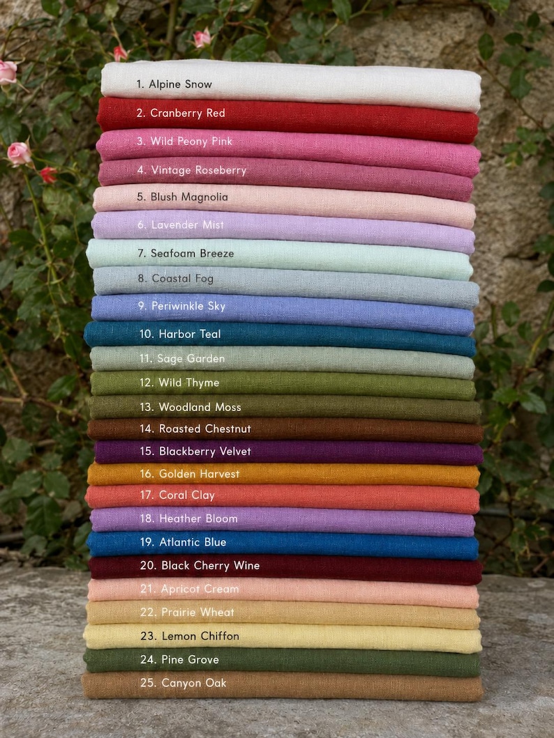 Op de afbeelding: Een stapel van vijfentwintig gevouwen stoffen stalen in verschillende kleuren, elk gelabeld met een nummer en naam, waaronder Alpine Snow, Cranberry Red, Wild Peony Pink, Vintage Roseberry, Blush Magnolia, Lavender Mist, Seafoam Breeze, Coastal Fog, Periwinkle Sky, Harbor Teal, Sage Garden, Wild Thyme, Woodland Moss, Roasted Chestnut, Blackberry Velvet, Golden Harvest, Coral Clay, Heather Bloom, Atlantic Blue, Black Cherry Wine, Apricot Cream, Prairie Wheat, Lemon Chiffon, Pine Grove en Canyon Oak.