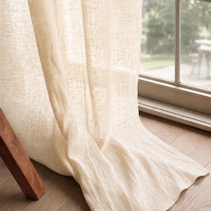 May include: A floor-length, semi-sheer curtain panel in a light beige, natural tone. The fabric features a visible, textured weave that allows soft light to filter through. The curtain hangs in loose, vertical folds, pooling slightly at the bottom.