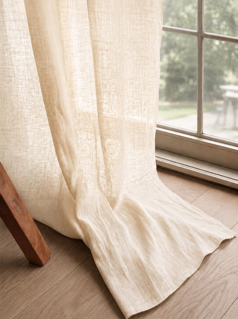 May include: A floor-length, semi-sheer curtain panel in a light beige, natural tone. The fabric features a visible, textured weave that allows soft light to filter through. The curtain hangs in loose, vertical folds, pooling slightly at the bottom.