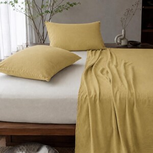 Op de afbeelding: Een bedset bestaande uit twee mosterdgele rechthoekige kussenslopen en een bijpassend mosterdgeel laken over een wit hoeslaken op een houten bedframe, met een grijs-witte kat die op de vloer naast het bed ligt te rusten.