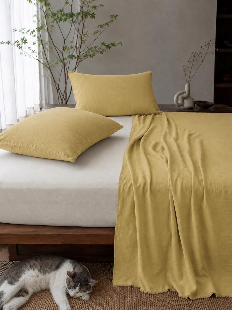 Op de afbeelding: Een bedset bestaande uit twee mosterdgele rechthoekige kussenslopen en een bijpassend mosterdgeel laken over een wit hoeslaken op een houten bedframe, met een grijs-witte kat die op de vloer naast het bed ligt te rusten.