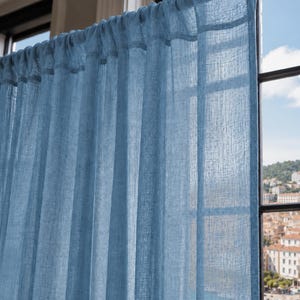 May include: A light blue, semi-sheer curtain panel with a visible woven texture hanging from a dark rod. The fabric is gathered at the top, creating vertical folds that allow light to pass through the material, highlighting the weave pattern.