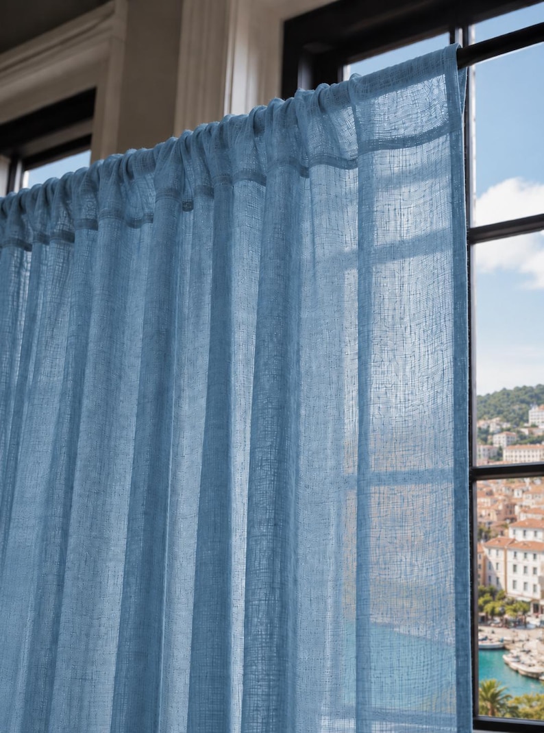 May include: A light blue, semi-sheer curtain panel with a visible woven texture hanging from a dark rod. The fabric is gathered at the top, creating vertical folds that allow light to pass through the material, highlighting the weave pattern.
