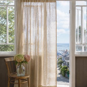 May include: A semi-sheer, light beige linen curtain hangs from a white rod over a doorway. The fabric has a visible, textured weave that allows soft light to filter through. The curtain is positioned next to a wooden chair holding a glass vase.