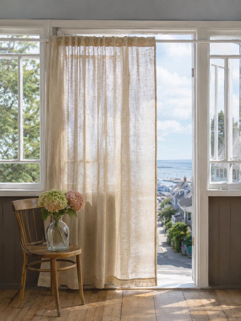 May include: A semi-sheer, light beige linen curtain hangs from a white rod over a doorway. The fabric has a visible, textured weave that allows soft light to filter through. The curtain is positioned next to a wooden chair holding a glass vase.