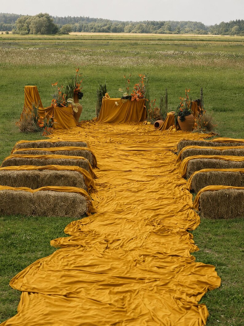 Op de afbeelding: Een buitenopstelling met rijen hooibalen bedekt met mosterdgele stof, die leiden naar een centrale tafel met een bijpassend kleed. Het arrangement bevat terracotta potten gevuld met oranje bloemen en hoge groene grassen.