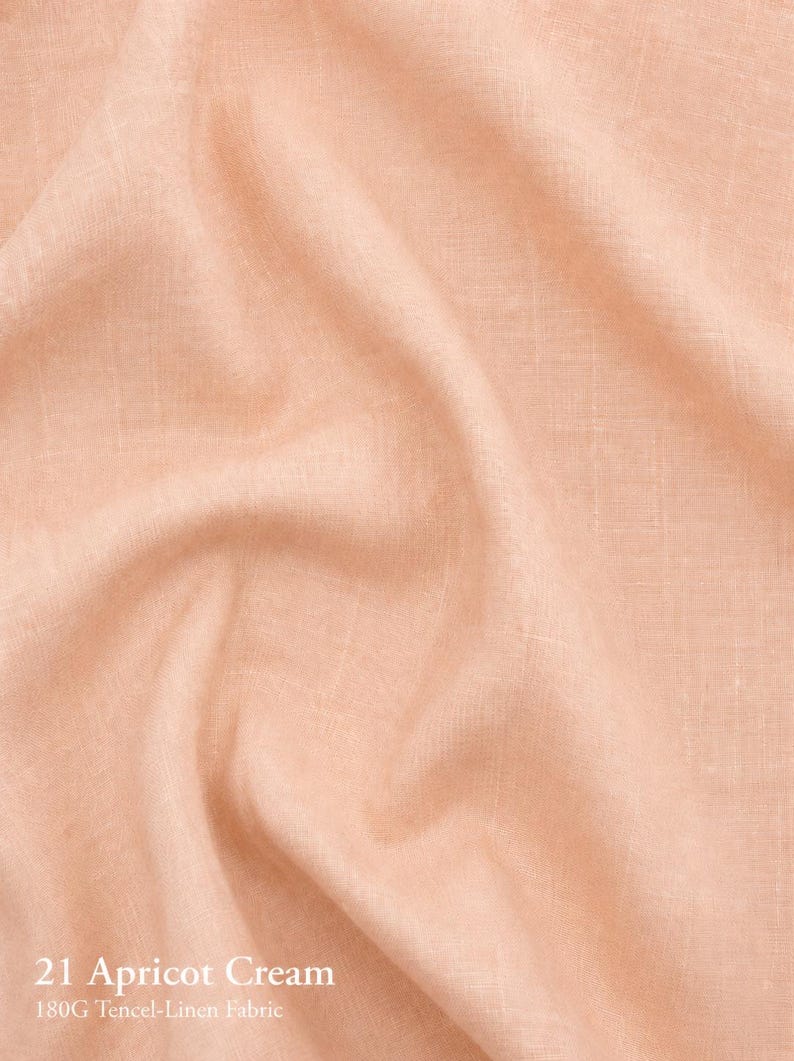 Puede incluir: Una pieza de tela color crema albaricoque con una textura tejida visible y pliegues suaves. El texto 21 Apricot Cream 180G Tencel-Linen Fabric est&aacute; impreso en blanco en la esquina inferior izquierda, indicando la composici&oacute;n del material.
