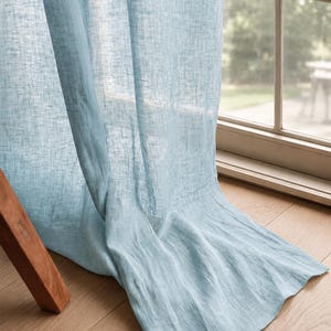 May include: A light blue curtain panel with a semi-sheer, textured weave. The fabric hangs in soft, vertical folds, extending from the top of the window frame down to the floor, creating a draped effect across the bottom edge of the window.