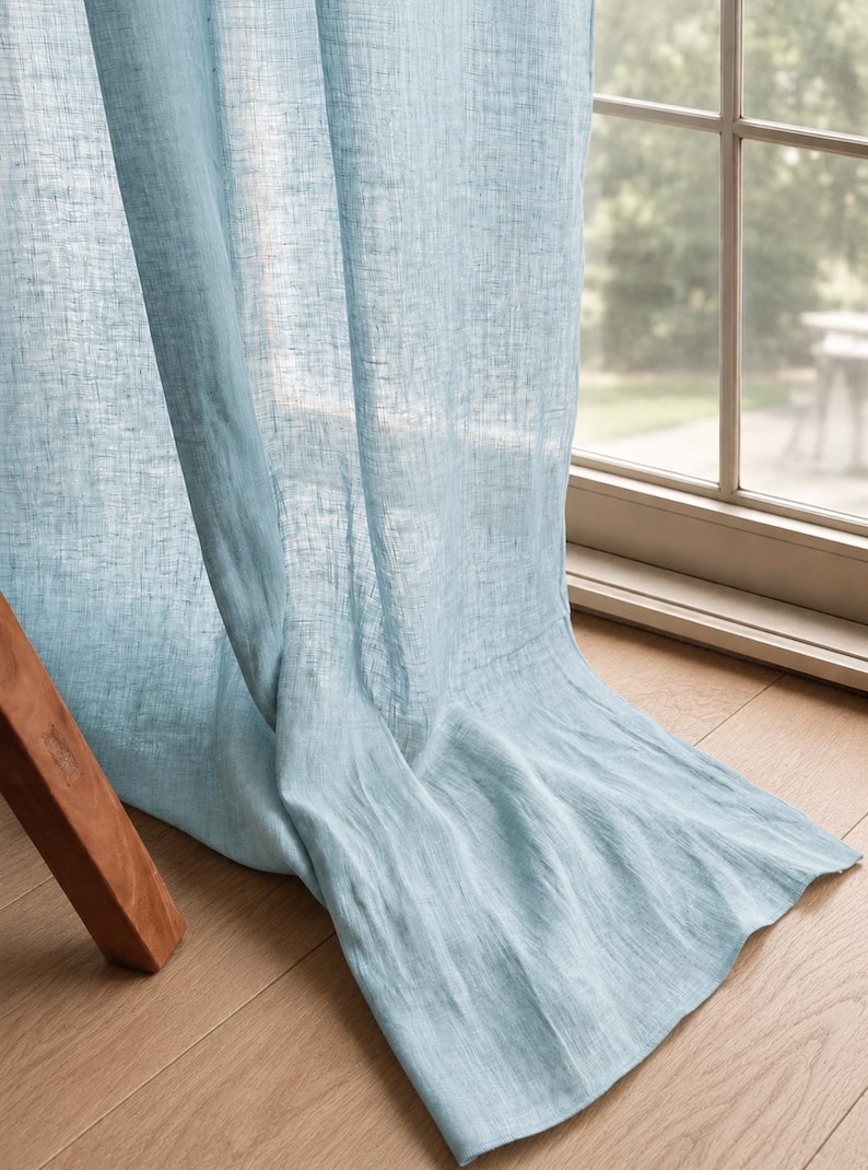 May include: A light blue curtain panel with a semi-sheer, textured weave. The fabric hangs in soft, vertical folds, extending from the top of the window frame down to the floor, creating a draped effect across the bottom edge of the window.