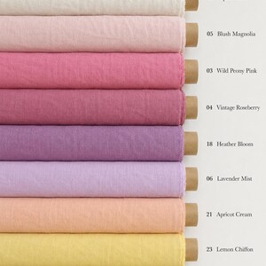 Op de afbeelding: Negen gestapelde rollen stof in verschillende kleuren met kartonnen kernen. De kleuren zijn gelabeld: 01 Alpine Snow, 05 Blush Magnolia, 03 Wild Peony Pink, 04 Vintage Roseberry, 18 Heather Bloom, 06 Lavender Mist, 21 Apricot Cream, 23 Lemon Chiffon, 22 Prairie Wheat.