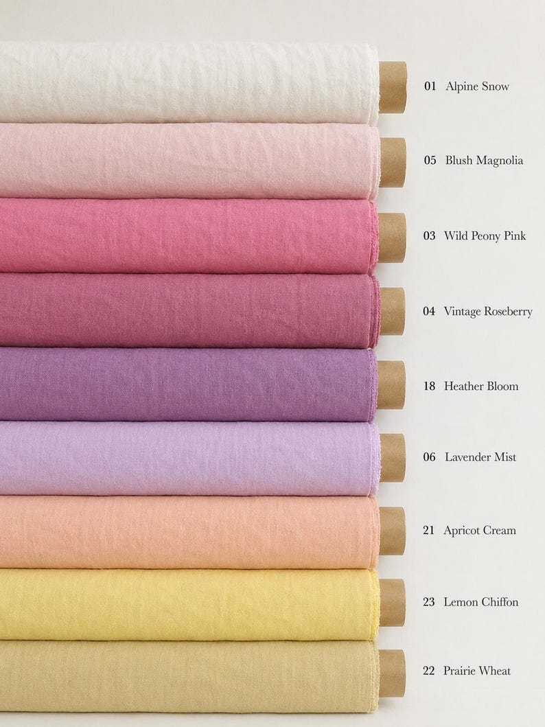 Op de afbeelding: Negen gestapelde rollen stof in verschillende kleuren met kartonnen kernen. De kleuren zijn gelabeld: 01 Alpine Snow, 05 Blush Magnolia, 03 Wild Peony Pink, 04 Vintage Roseberry, 18 Heather Bloom, 06 Lavender Mist, 21 Apricot Cream, 23 Lemon Chiffon, 22 Prairie Wheat.