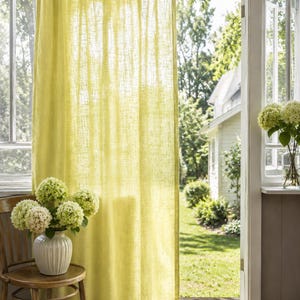 May include: A sheer, light yellow curtain with a visible woven texture hangs from a rod. The fabric is semi-transparent, allowing natural light to filter through, and features a simple rod pocket header at the top and a straight hem at the bottom.