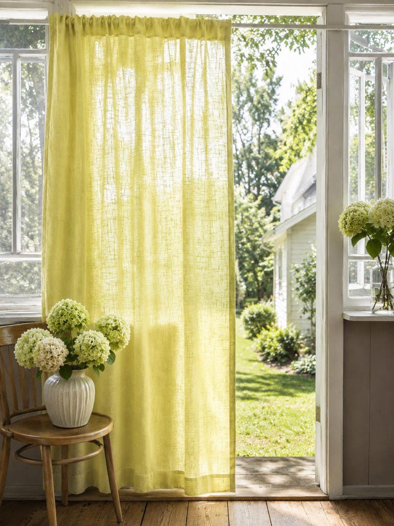 May include: A sheer, light yellow curtain with a visible woven texture hangs from a rod. The fabric is semi-transparent, allowing natural light to filter through, and features a simple rod pocket header at the top and a straight hem at the bottom.