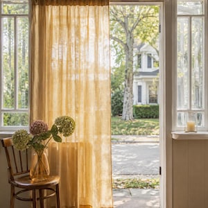 May include: A sheer, golden-yellow curtain panel hangs from a rod over a doorway. The fabric has a visible, slightly textured weave that allows light to pass through. The curtain is positioned next to a wooden chair holding a glass vase with flowers.