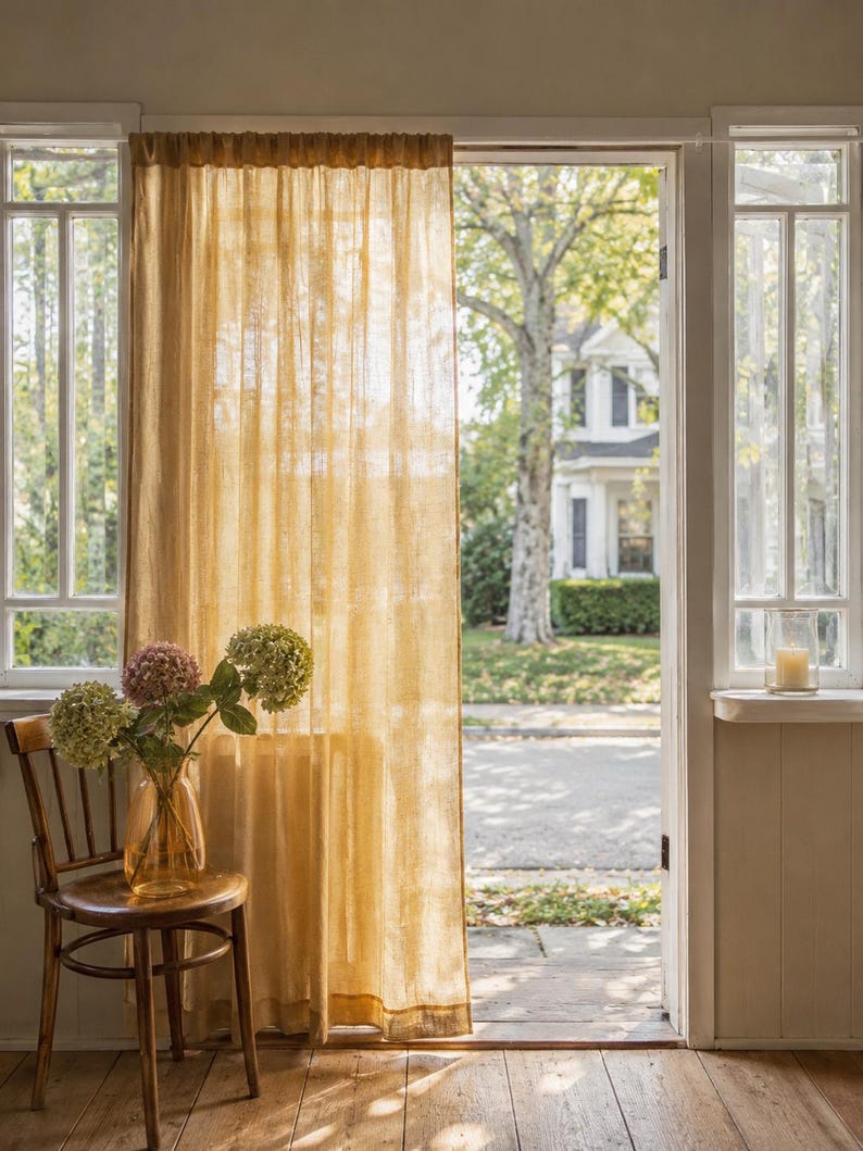 May include: A sheer, golden-yellow curtain panel hangs from a rod over a doorway. The fabric has a visible, slightly textured weave that allows light to pass through. The curtain is positioned next to a wooden chair holding a glass vase with flowers.
