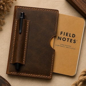Könnte beinhalten: Ein dunkelbrauner Notizbuchumschlag aus Leder mit sichtbaren Nähten und einer Stiftschlaufe, die einen schwarzen Stift hält. Ein beigefarbenes 48-seitiges Field Notes Notizbuch steckt darin, mit dem Text Field Notes, 48-Page Memo Book, Durable Materials, Made in the U.S.A.