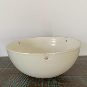 May include: Off-white, semi-spherical bowl with three small holes near the rim. The bowl is set against a white background and rests on a distressed, dark wood surface with teal accents. The bowl's smooth surface contrasts with the textured wood.