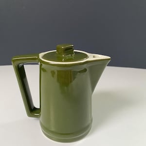 Könnte beinhalten: Kleine, olivgrüne Keramik-Kaffeekanne mit passendem Deckel und weißem Rand. Der Griff ist rechteckig und der Ausguss spitz. Die Kanne steht auf einer weißen Oberfläche mit dunklem blauem Hintergrund.
