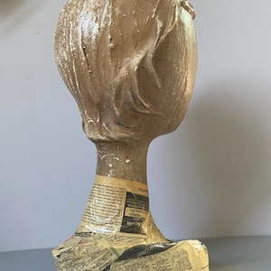 May include: A vintage mannequin head with a textured, light brown finish. The head features sculpted hair and is mounted on a neck and shoulder form wrapped in newspaper. Ideal for displaying wigs, hats, or jewelry.