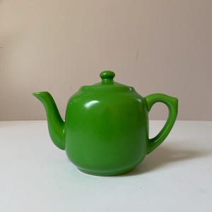 Könnte beinhalten: Eine leuchtend grüne Keramik-Teekanne mit rundem Körper, gebogenem Ausguss und Schlaufengriff. Der Deckel hat einen kleinen Knopf. Die Teekanne steht auf einer weißen Oberfläche.