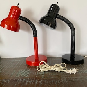 May include: Two desk lamps, one red and one black, with flexible black gooseneck arms. Each lamp has a round base and a small on/off switch on top. A white power cord with a plug is visible.