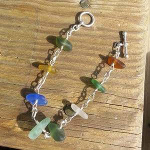 May include: A silver chain bracelet with a variety of sea glass beads in shades of green, blue, and yellow. The bracelet has a lobster clasp closure.